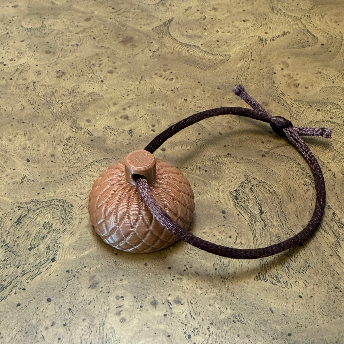 Small round textured cap for the acorn scent hide.  The cap is placed on a textured wood-grain surface and is showing the hanging variant that allows users to hang the cap as a suspended hide. 