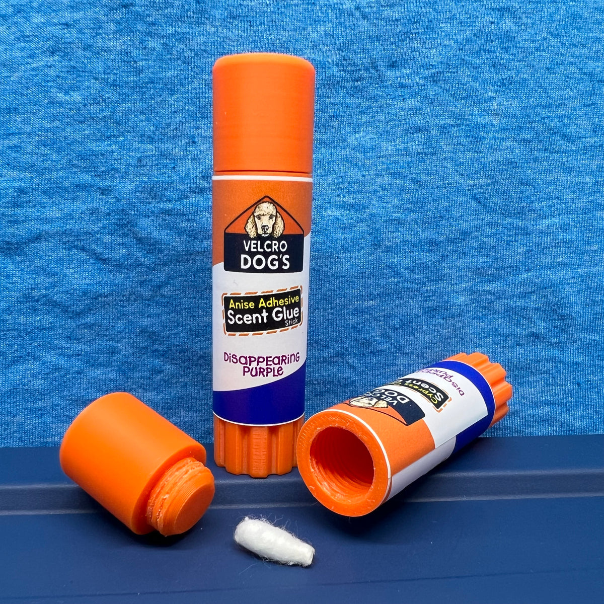 "Anise Adhesive" variant of the Velcro Dog's Super Cured Scent Glue Hide.  This fun training aid includes an orange squeeze tube with a custom label.  Shown with the cap off and a small Q-tip on a blue textured background.