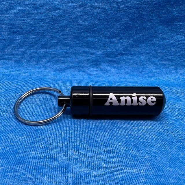 A black cylindrical keychain container labeled 'Anise' in white text, attached to a silver metal key ring, placed on a textured blue fabric background.