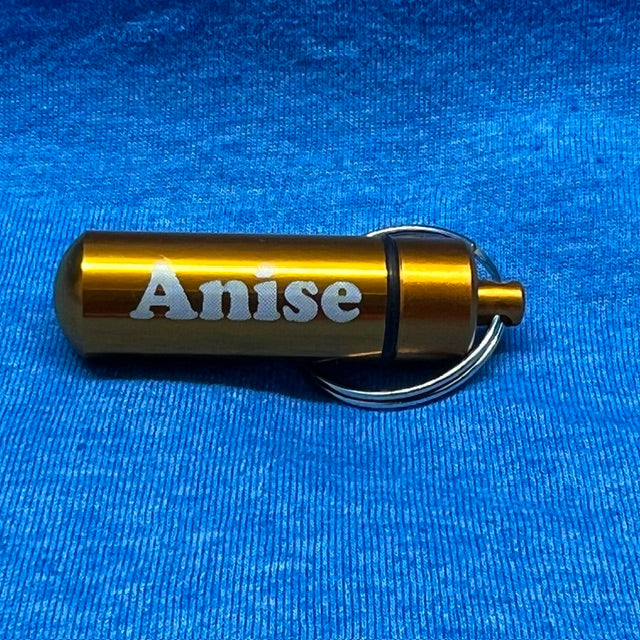 A small metallic gold cylindrical keychain container engraved with the name 'Anise' in white, attached to a silver metal key ring, resting on a textured blue fabric background.