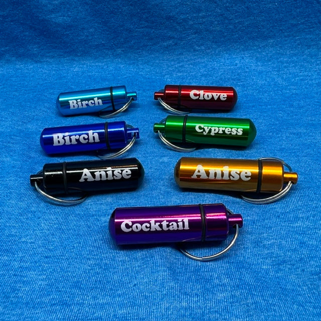 Seven small cylindrical metal keychain containers labeled Birch, Clove, Cypress, Anise, and Cocktail, shown in blue, red, green, black, gold, and purple on a textured blue fabric background.