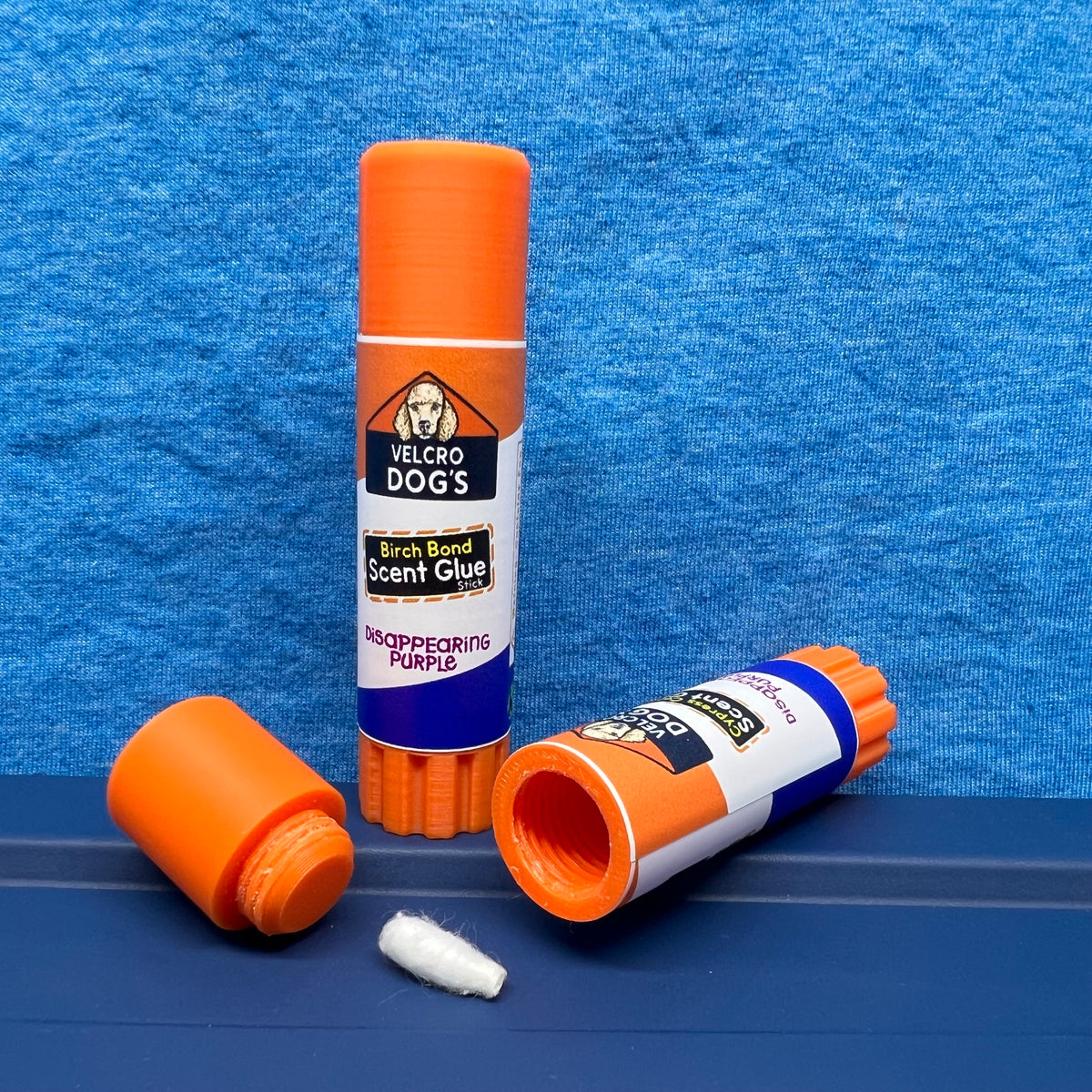"Birch Bond" variant of the Velcro Dog's Super Cured Scent Glue Hide.  This fun training aid includes an orange squeeze tube with a custom label.  Shown with the cap off and a small Q-tip on a blue textured background.