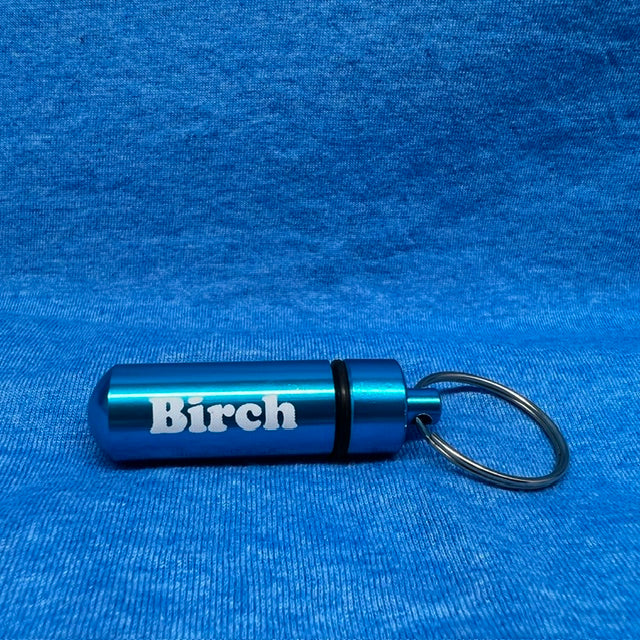 A metallic blue cylindrical keychain container engraved with the word 'Birch' in white, attached to a black split ring, resting on a textured blue fabric background.
