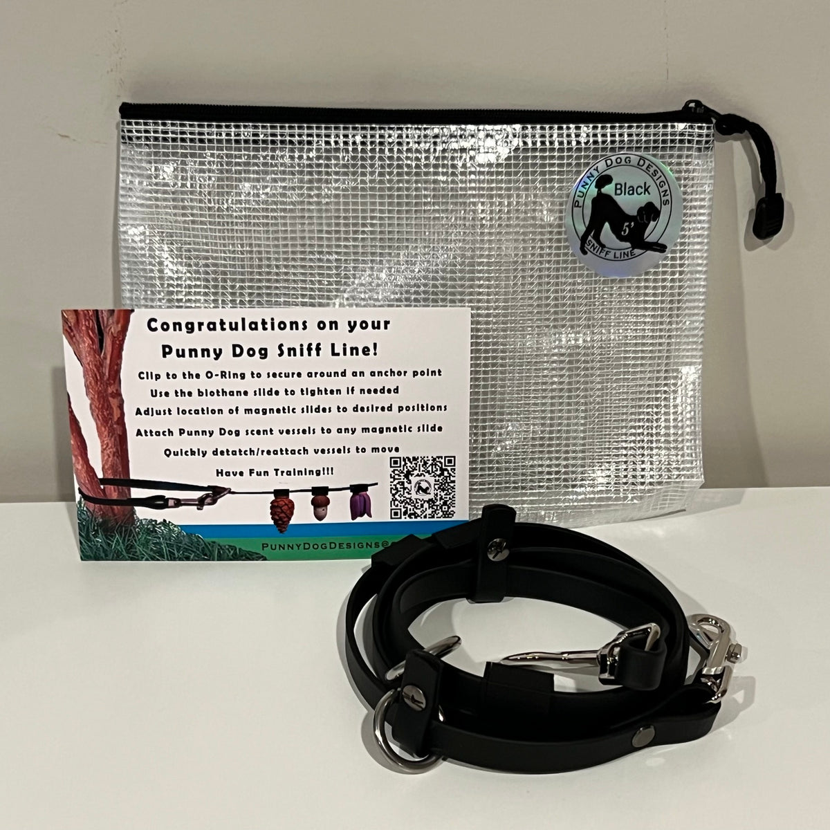 A dog training accessory set displayed on a white surface against a light gray wall, including a clear mesh zip pouch, a black biothane line that has a metal clip, and a printed instruction card.
