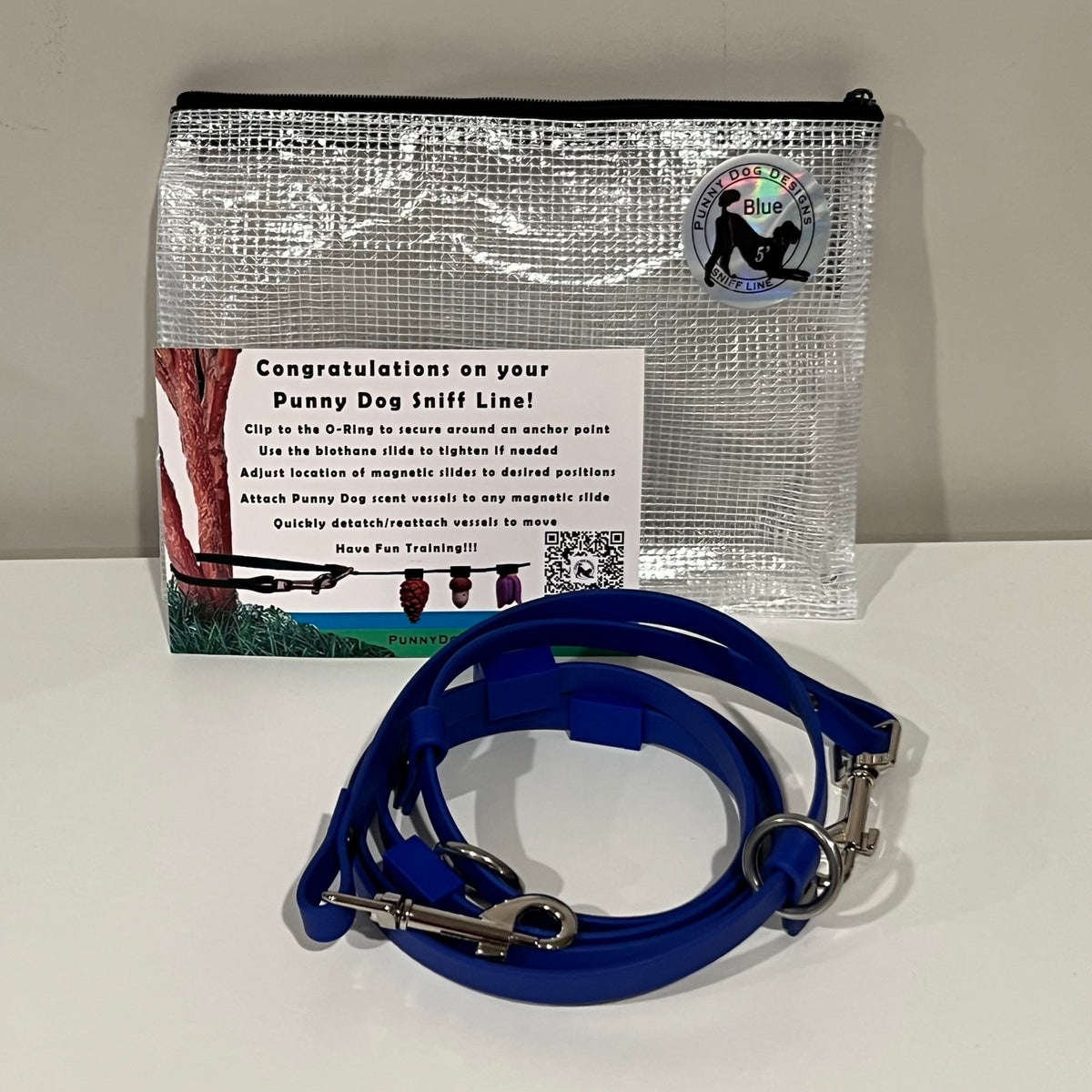 A dog training accessory set displayed on a white surface against a light gray wall, including a clear mesh zip pouch, a blue biothane line that has a metal clip, and a printed instruction card.