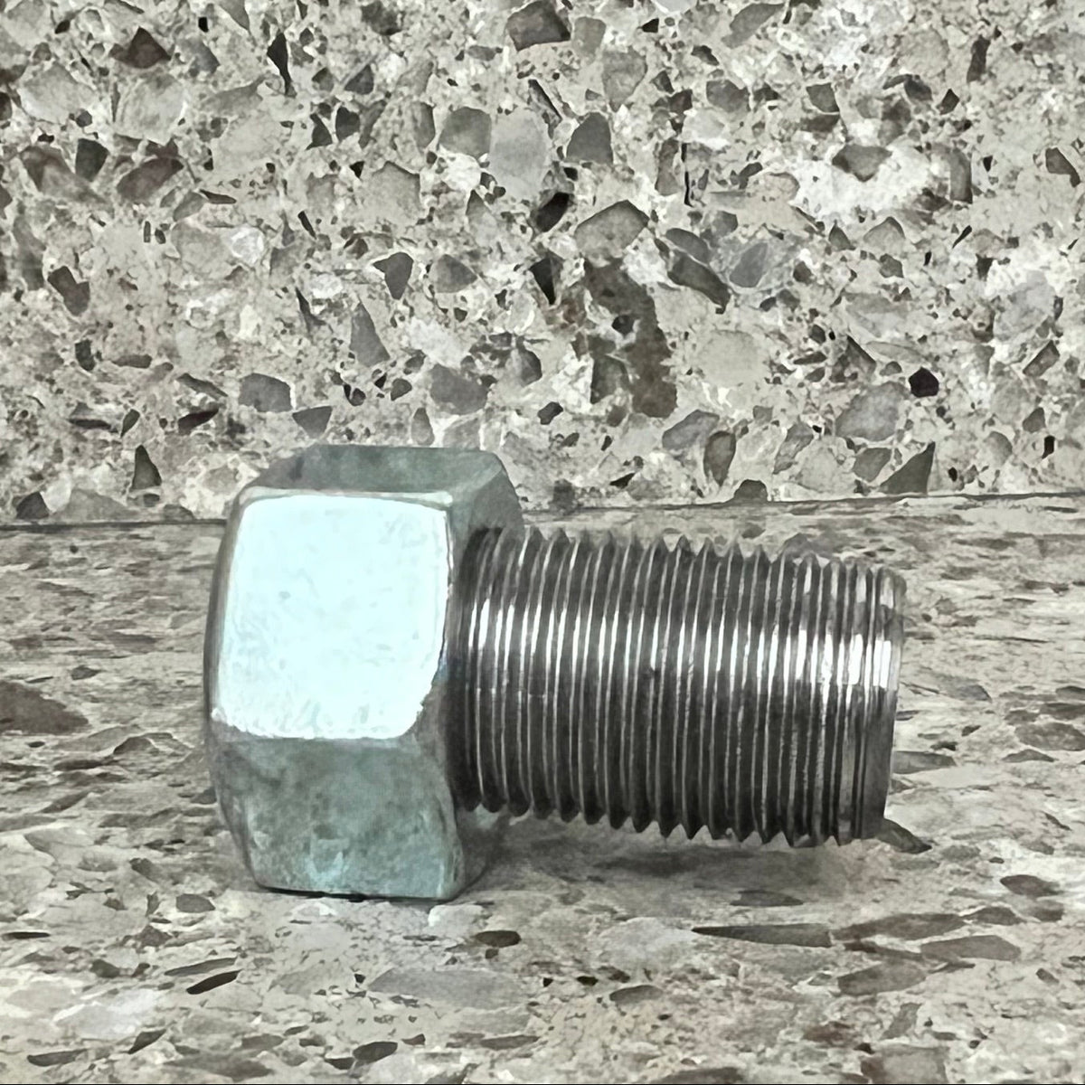 Scent work hide designed to look like a single metal threaded bolt or pipe fitting with a hexagonal head, photographed laying on its side on a speckled stone surface against a matching stone background.