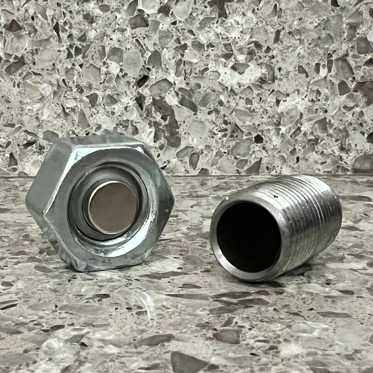 Scent work hide designed to look like a metal bolt.   photographed laying on its side open revealing that the head is really a nut with a magnet and the shaft is hollow.  Photographed on a speckled stone surface against a matching stone background. 