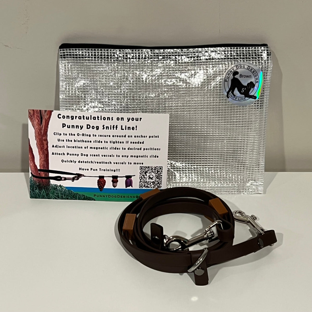 A dog training accessory set displayed on a white surface against a light gray wall, including a clear mesh zip pouch, a brown biothane line that has a metal clip, and a printed instruction card.