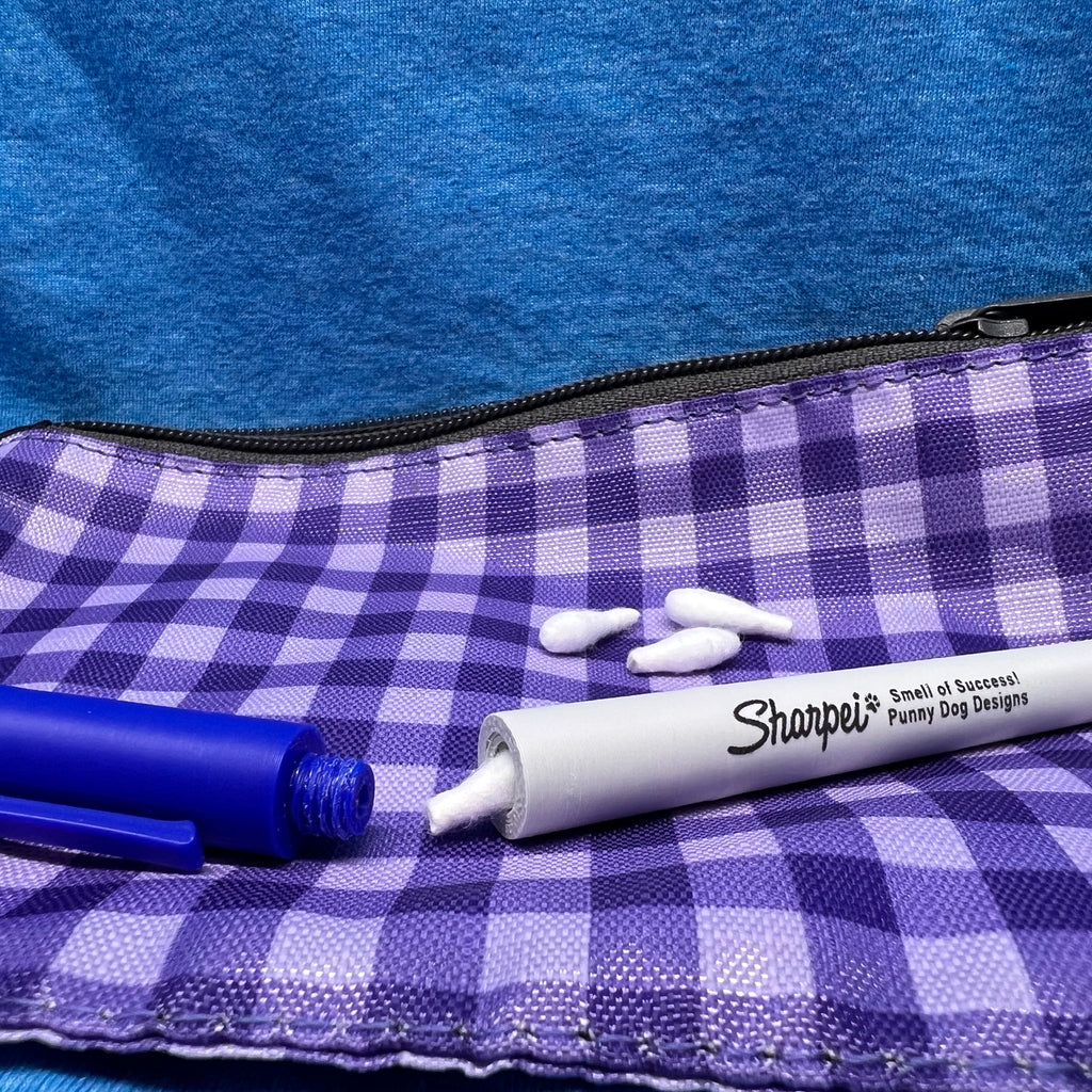 A close-up of a single blue marker hide.  The cap is off showing that it attaches securely using a threaded screw mechanism.  A Q-Tip is partially out of the body of the hide.  The Image is set on purple and white gingham fabric pouch with a black zipper on a blue textured background.