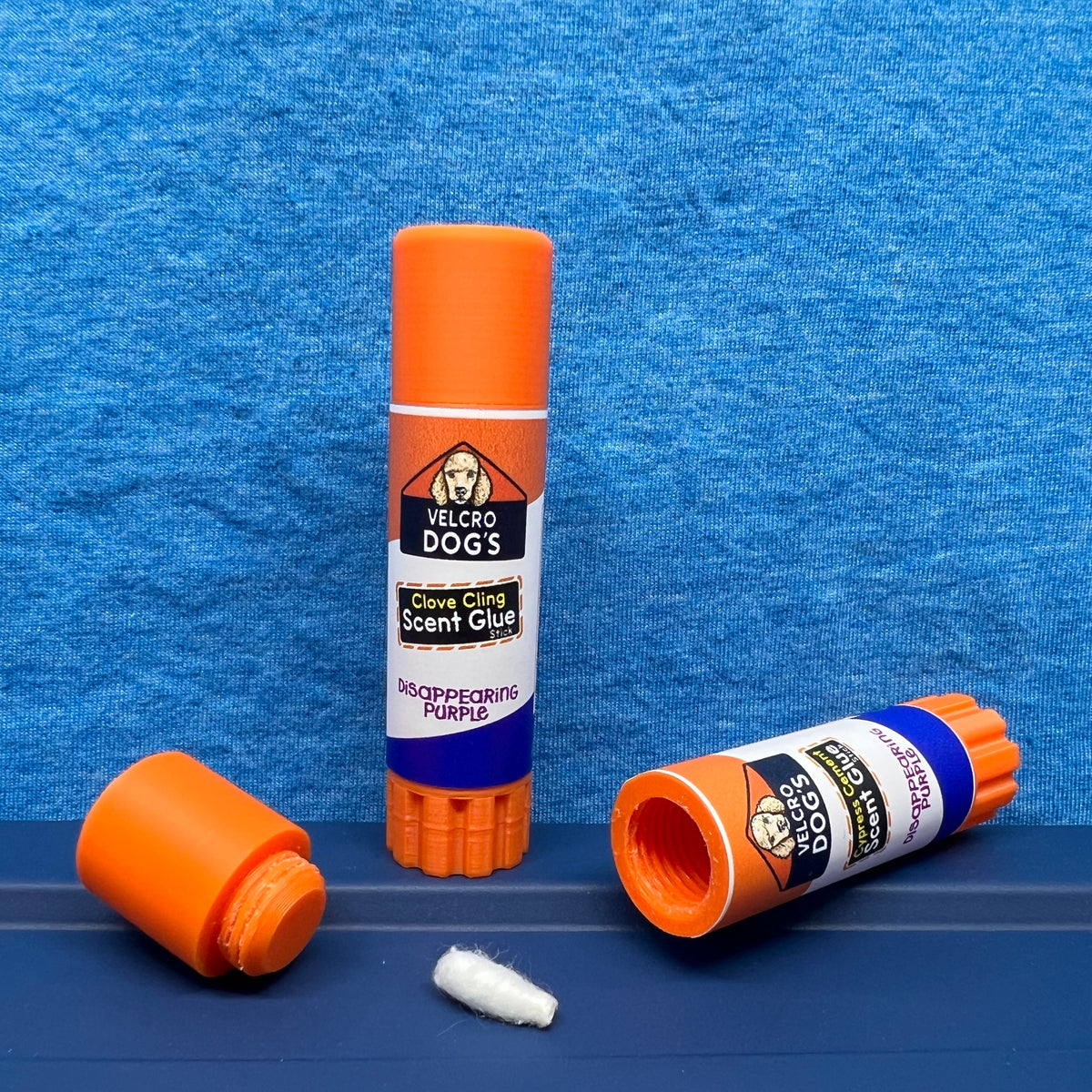 "Clove Cling" variant of the Velcro Dog's Super Cured Scent Glue Hide.  This fun training aid includes an orange squeeze tube with a custom label.  Shown with the cap off and a small Q-tip on a blue textured background.