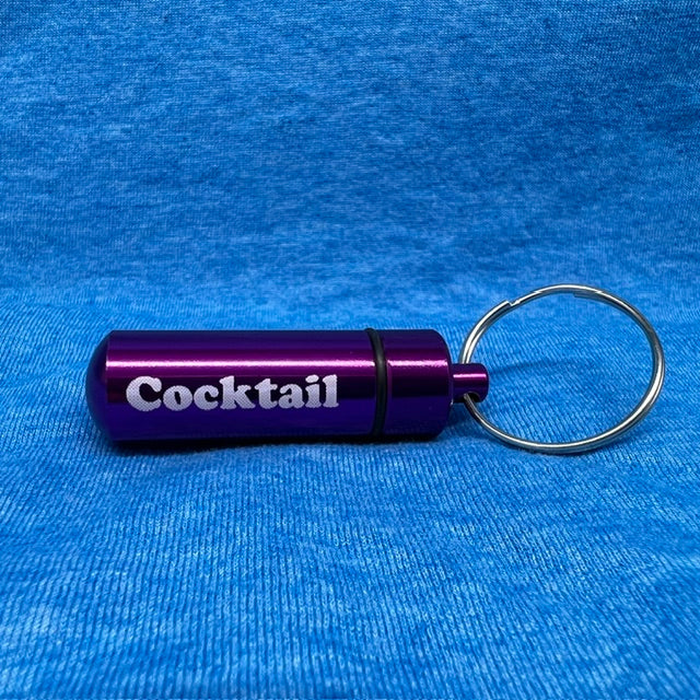 A purple anodized metal cocktail keychain featuring the word 'Cocktail' in white text, complete with a silver split ring, resting on a textured blue fabric background.