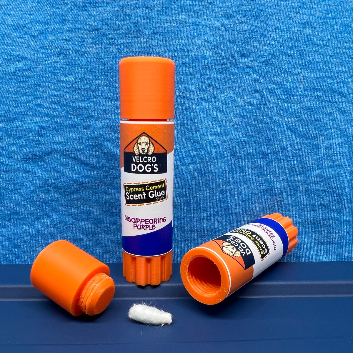 "Cypress Cement" variant of the Velcro Dog's Super Cured Scent Glue Hide.  This fun training aid includes an orange squeeze tube with a custom label.  Shown with the cap off and a small Q-tip on a blue textured background.