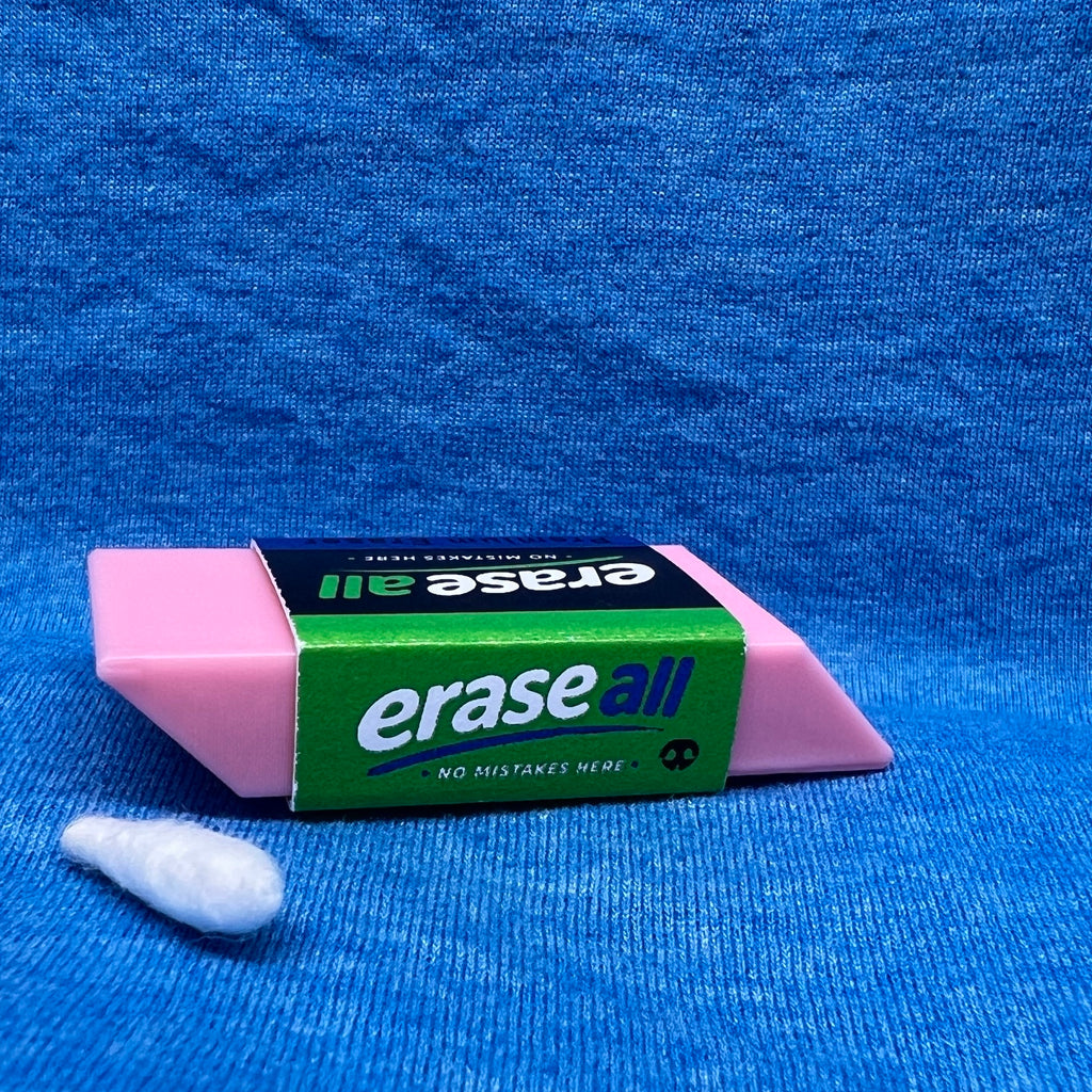 Clever scent work hide disguised as a  pink eraser.  The packaging band is green, black and blue.  The hide is shown placed on a textured blue fabric background next to a small white Q-tip used for scent work.