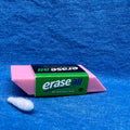 Clever scent work hide disguised as a  pink eraser.  The packaging band is green, black and blue.  The hide is shown placed on a textured blue fabric background next to a small white Q-tip used for scent work.