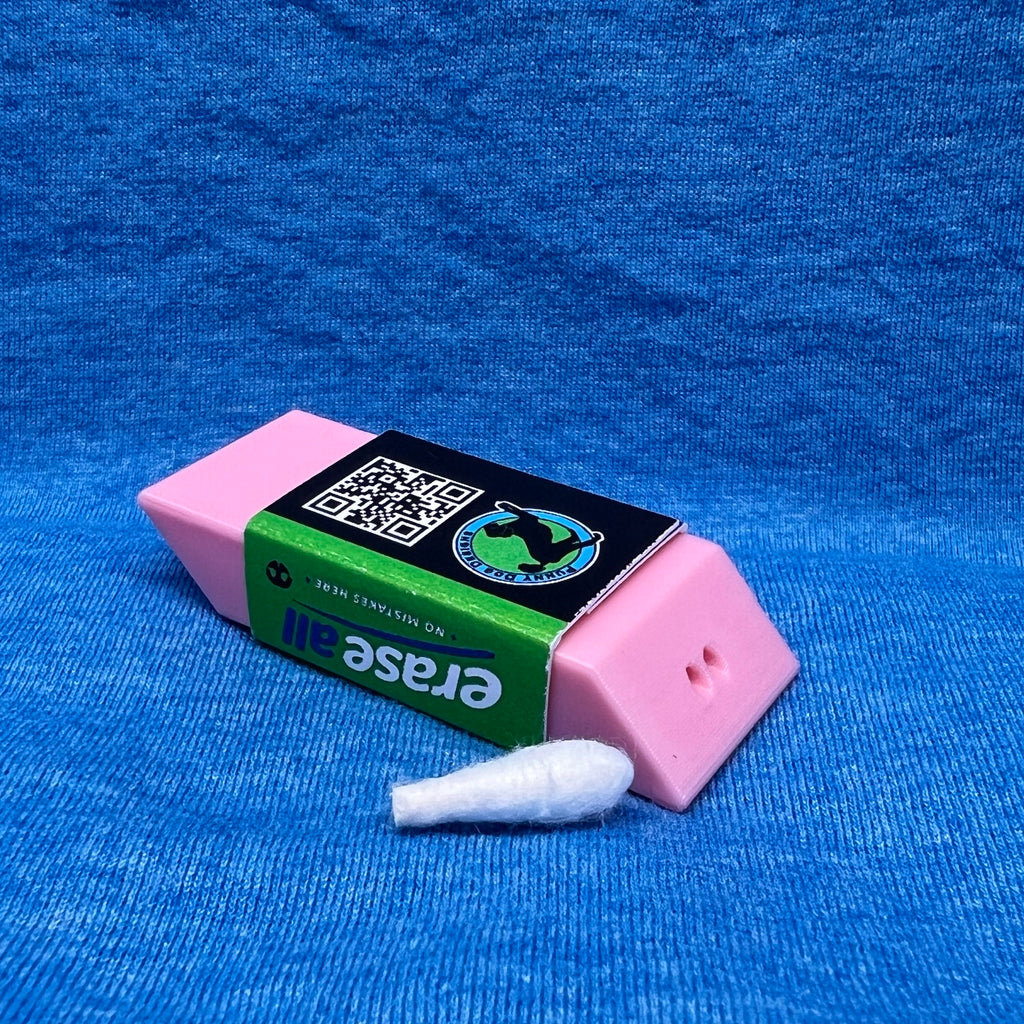 The eraser scent work hide shown upside down.  From this angle you can see what appears to be a pink eraser, but it has two holes at one end for the odor to escape.   The packaging band is green, black and blue and on the bottom shows a QR code and the Punny Dog brand logo.  The hide is shown on a textured blue fabric background next to a small white Q-tip used for scent work.