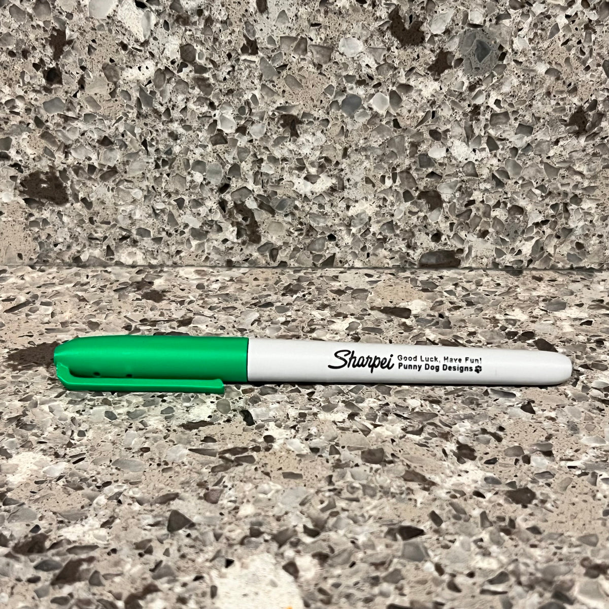 A green and white Sharpie marker shaped scent work hide lying on a speckled stone countertop with a matching stone backsplash behind it.