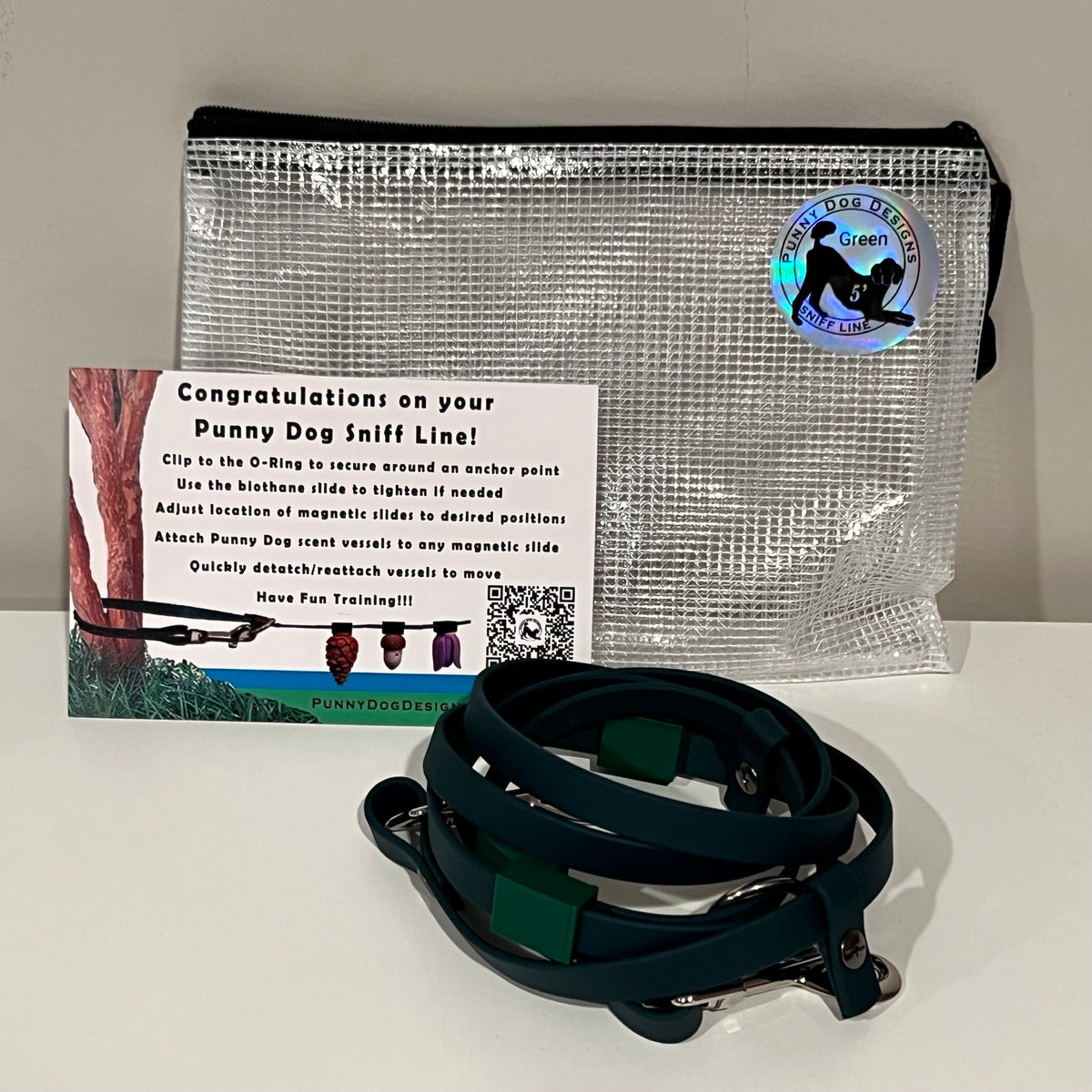 A dog training accessory set displayed on a white surface against a light gray wall, including a clear mesh zip pouch, a dark green biothane line that has a metal clip, and a printed instruction card.
