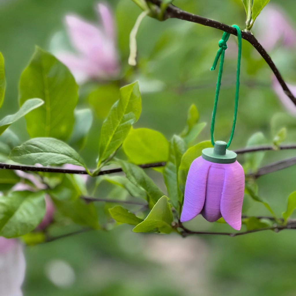 A tulip shaped scent work hide shown hanging from a green top and green hanging cord.  Suspended from a tree branch with green leaves and soft pink blossoms in the background.