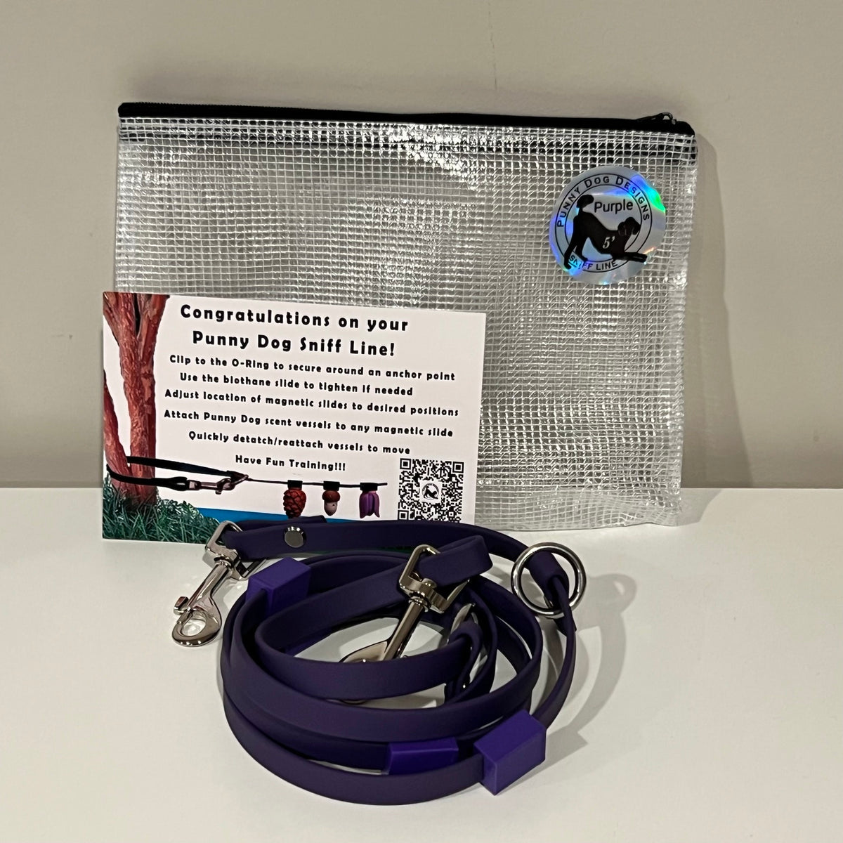A dog training accessory set displayed on a white surface against a light gray wall, including a clear mesh zip pouch, a pruple biothane line that has a metal clip, and a printed instruction card.