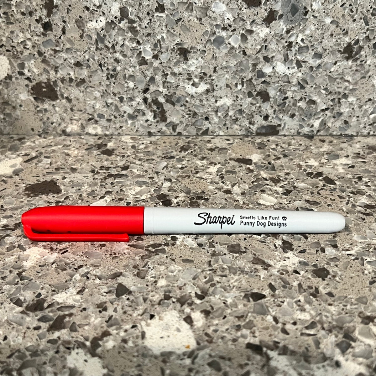 A red and white Sharpie marker shaped scent work hide lying on a speckled stone countertop with a matching stone backsplash behind it.