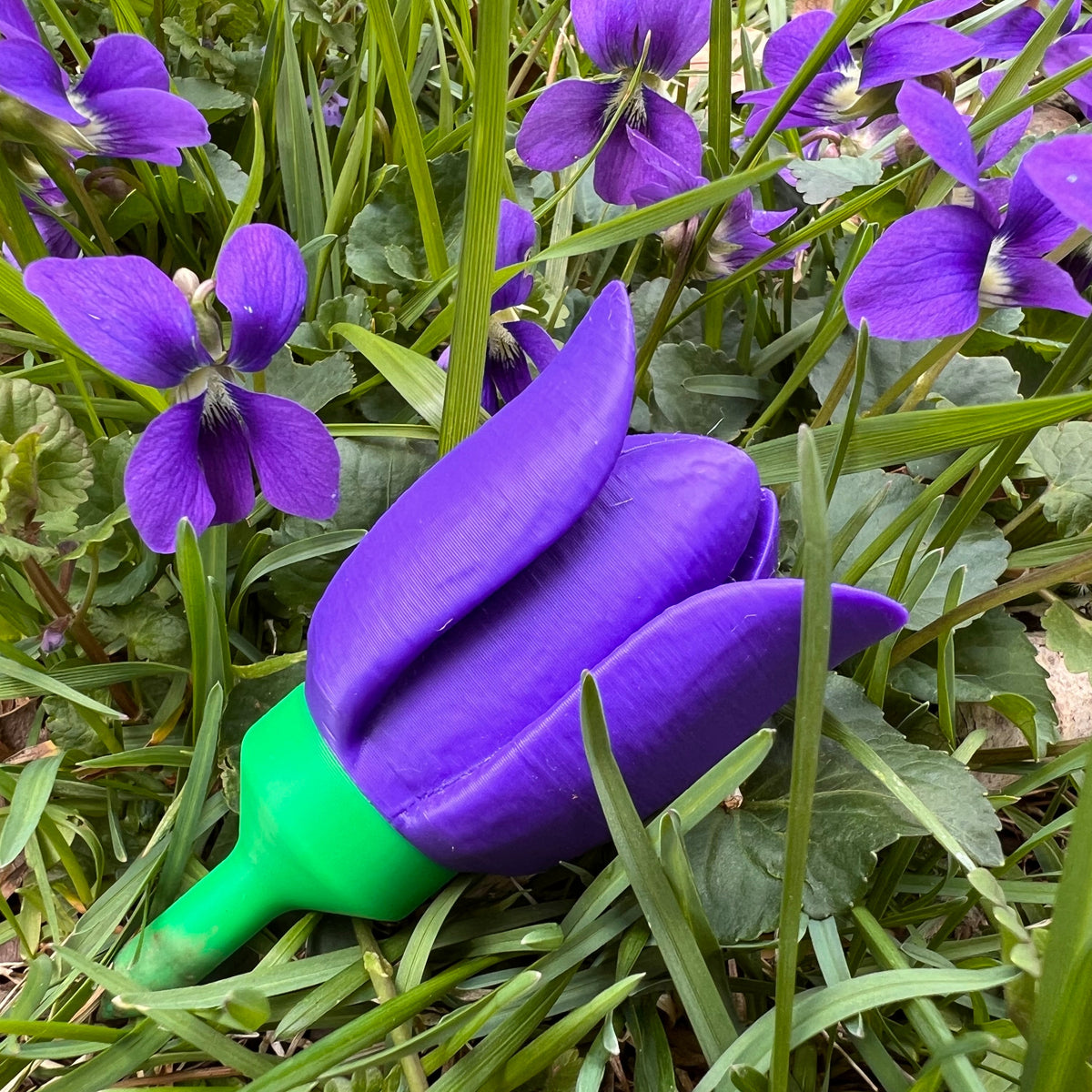 A purple tulip-shaped novelty item with a green stem lying on green grass among small purple flowers.