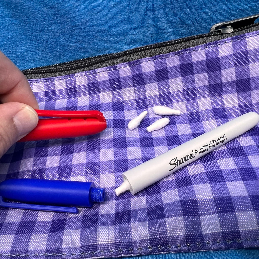 A close-up of a two marker hides (one blue & one red).  The red cap is held by a hand lifting the clip slightly from the body of the cap revealing two holes for odor to escape under the clip.  

The blue marker hide is in the foreground and the cap is off showing that it attaches securely using a threaded screw mechanism.  A Q-Tip is partially out of the body of the hide.  

The Image is set on purple and white gingham fabric pouch with a black zipper on a blue textured background.