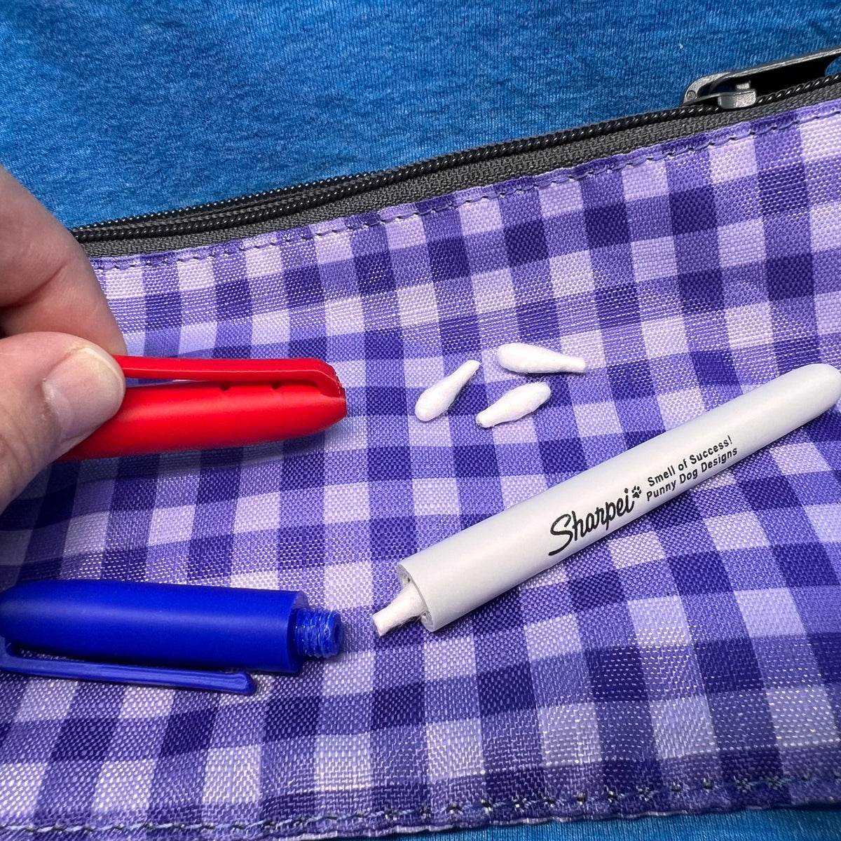 A close-up of a two marker hides (one blue & one red).  The red cap is held by a hand lifting the clip slightly from the body of the cap revealing two holes for odor to escape under the clip.  

The blue marker hide is in the foreground and the cap is off showing that it attaches securely using a threaded screw mechanism.  A Q-Tip is partially out of the body of the hide.  

The Image is set on purple and white gingham fabric pouch with a black zipper on a blue textured background.