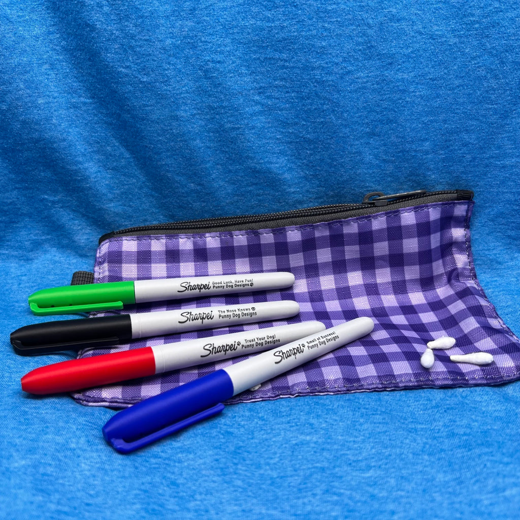 Image showing four cleverly disguised scent work hides that appear to be Sharpie Markers in green, black, red, and blue.  Hides are placed on a small zippered pouch with a purple and white gingham pattern set against a textured blue fabric background.