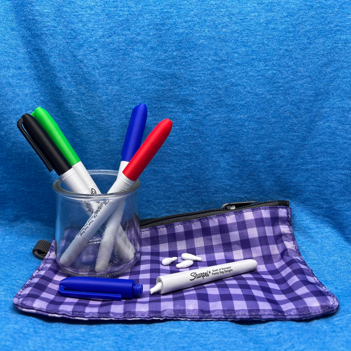 A purple and white gingham fabric pencil pouch with a black zipper, shown with several scent work hides that appear to be markers inside a clear glass cup.  A single loose marker hide is open separating the cap from the body of the hide with a Q-Tip halfway out.  The cap fastens to the sharpie using a threaded cap.  Image is set on a bright blue textured fabric background. 
