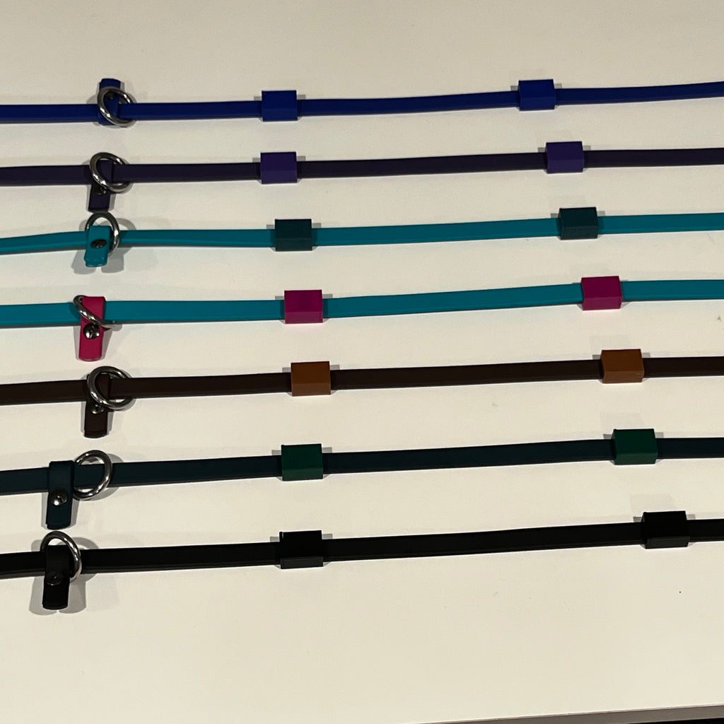Seven adjustable dog training aids called "Sniff Lines" in different colors (blue, purple, teal, brown, green, and black) arranged horizontally on a light surface. Each sniff lines features a metal ring and a sliding adjustment tab, and a small square magnetic "slider" in a complimentary color used to attach scent work vessels. 