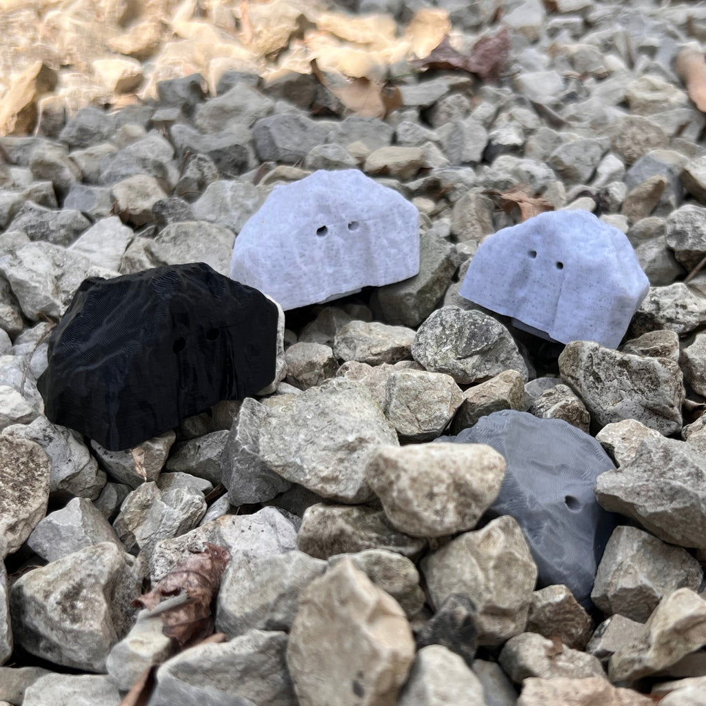 Four small stones shaped scent work hides in black, light marble gray, medium gray.  They are arranged on a bed of gray gravel outdoors.