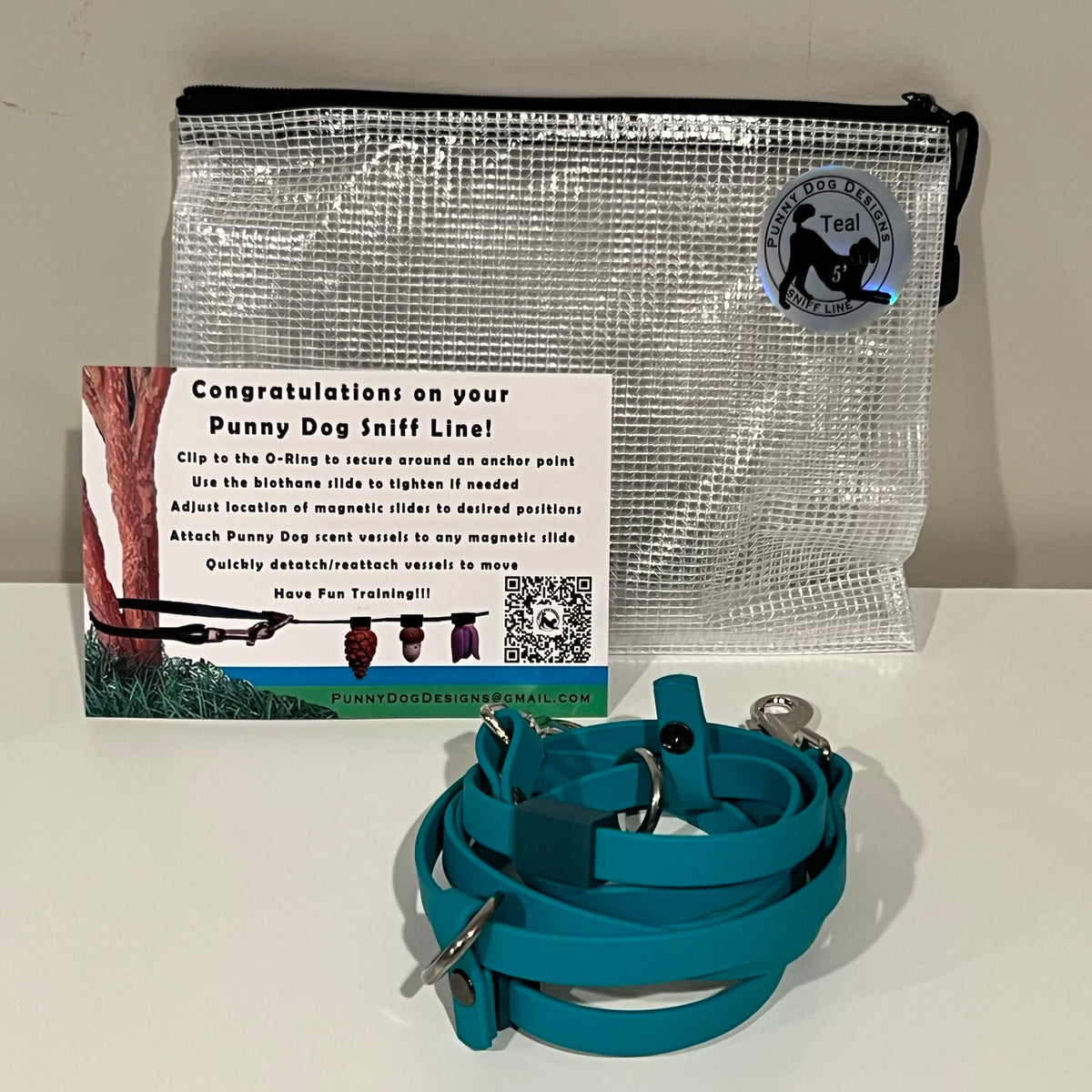 A dog training accessory set displayed on a white surface against a light gray wall, including a clear mesh zip pouch, a teal biothane line that has a metal clip, and a printed instruction card.