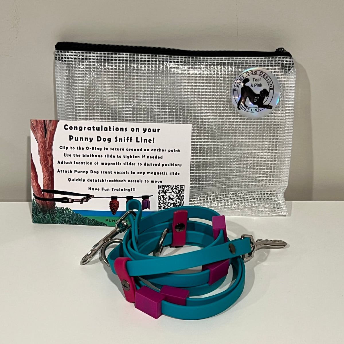 A dog training accessory set displayed on a white surface against a light gray wall, including a clear mesh zip pouch, a teal biothane line that has a metal clip, and a printed instruction card.