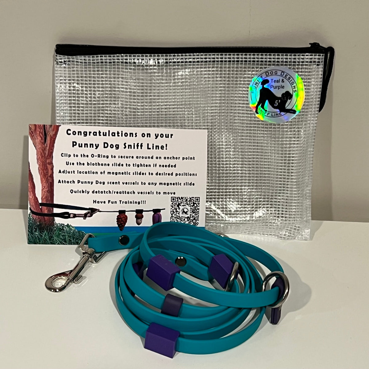 A dog training accessory set displayed on a white surface against a light gray wall, including a clear mesh zip pouch, a teal biothane line that has a metal clip, and a printed instruction card.