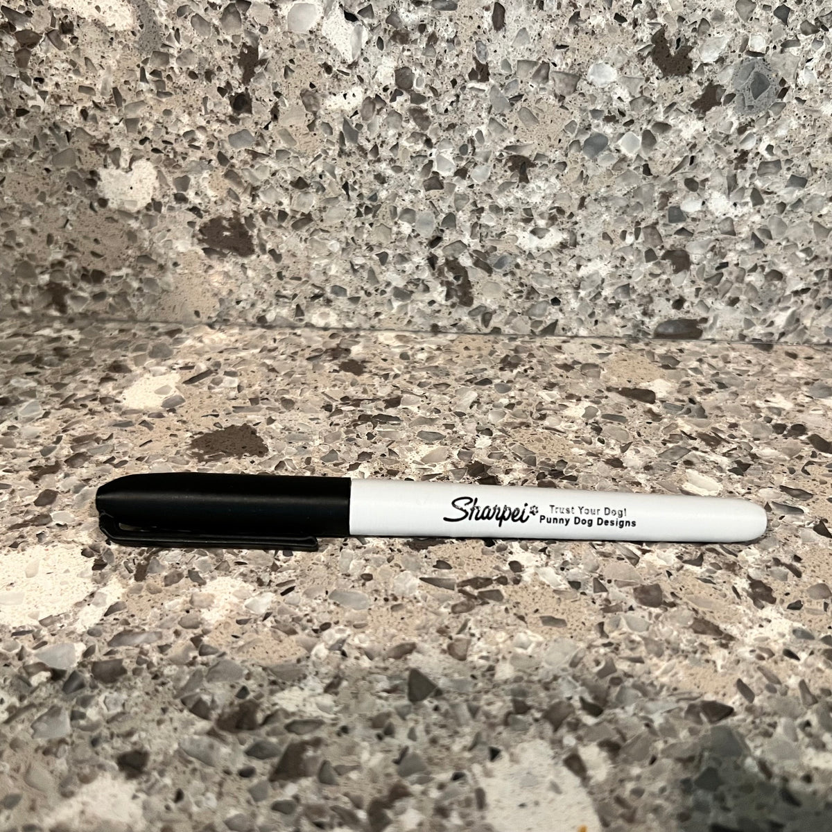 A black and white Sharpie marker shaped scent work hide lying on a speckled stone countertop with a matching stone backsplash behind it.