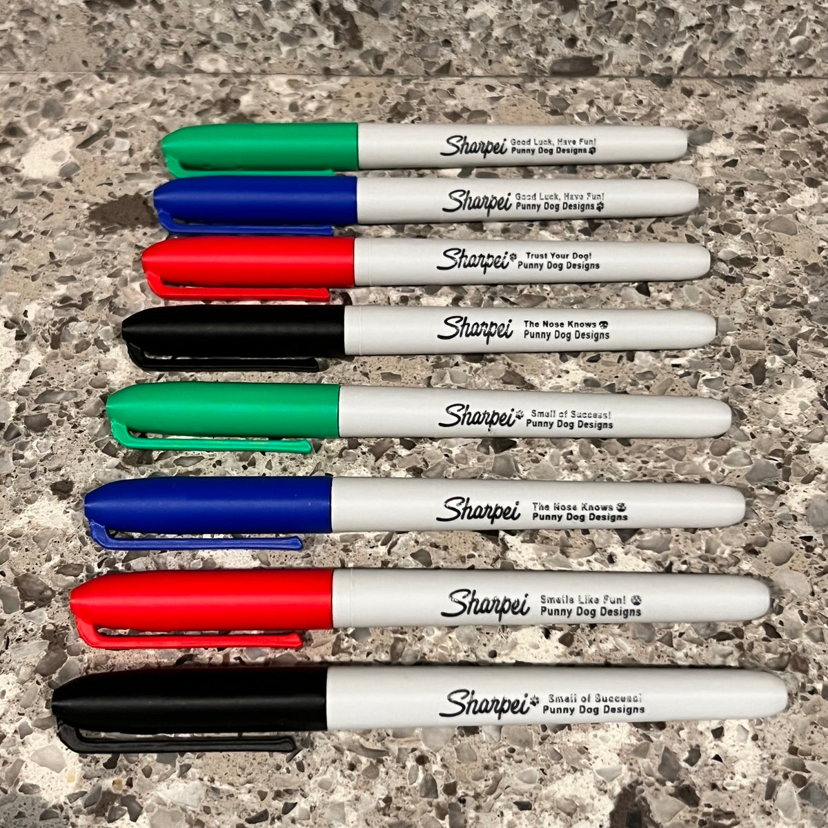 Seven scent work hides shaped like Sharpie permanent marker pens in assorted colors are lined up vertically on a speckled stone surface. The markers have light grey barrels with black printed text and caps in green, blue, red, and black.